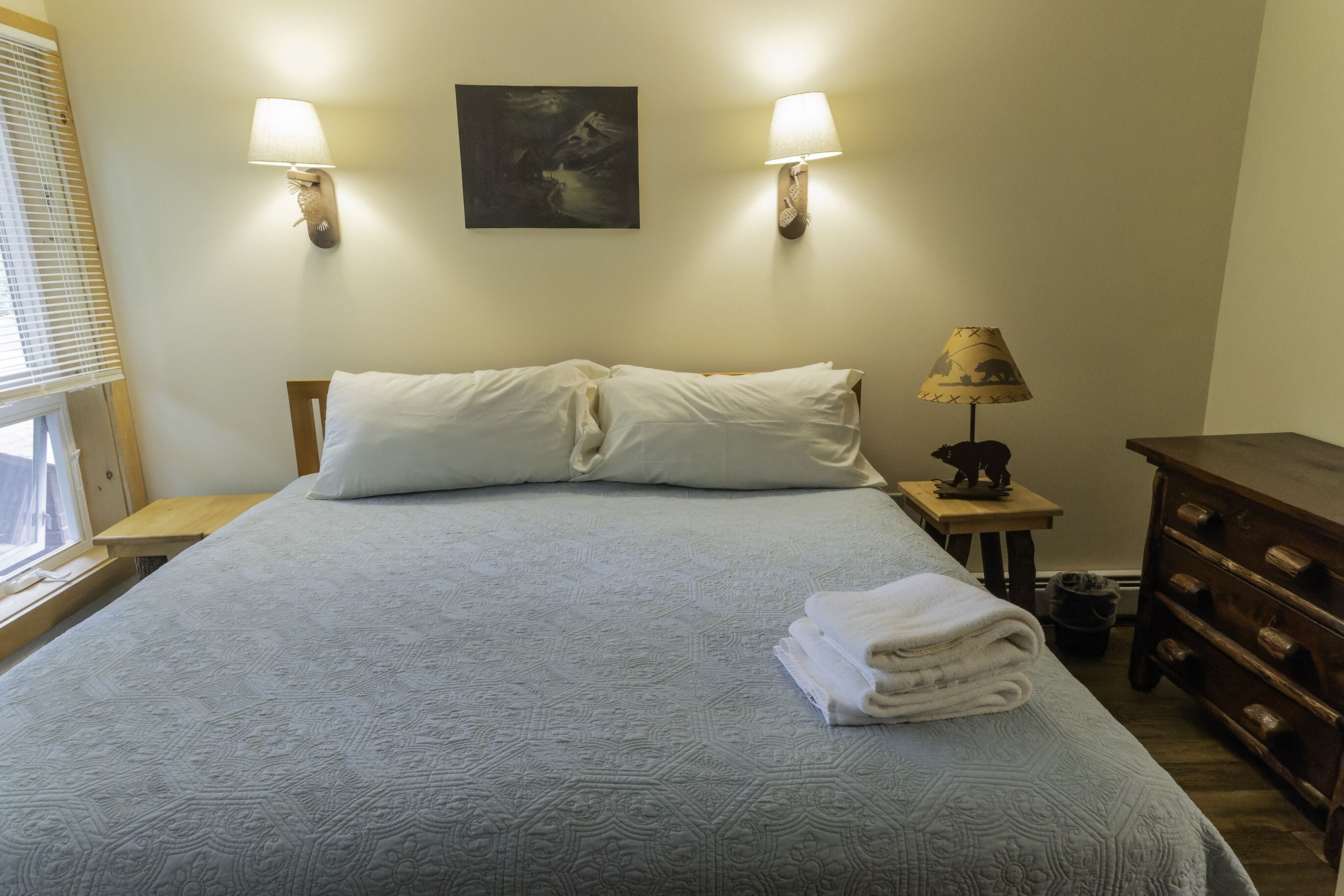 Una cama bien hecha con una colcha estampada, dos lámparas de pared, una mesita de noche con lámpara y un aparador de madera.