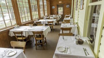 A dining area with multiple tables set for meals, wooden chairs, and large windows providing natural light.