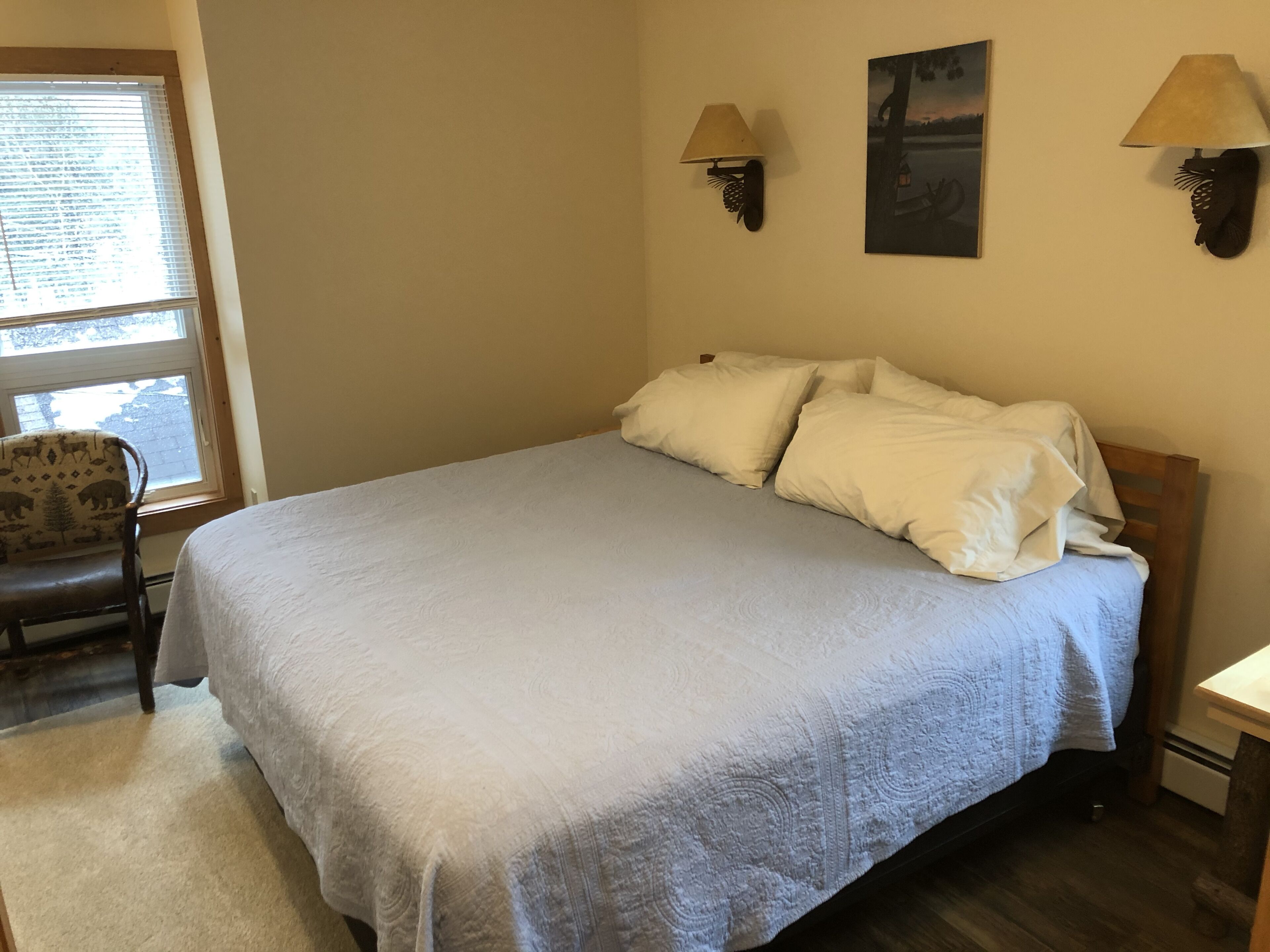 A bedroom with a bed, two wall-mounted lamps, a chair, a window, and a painting on the wall.