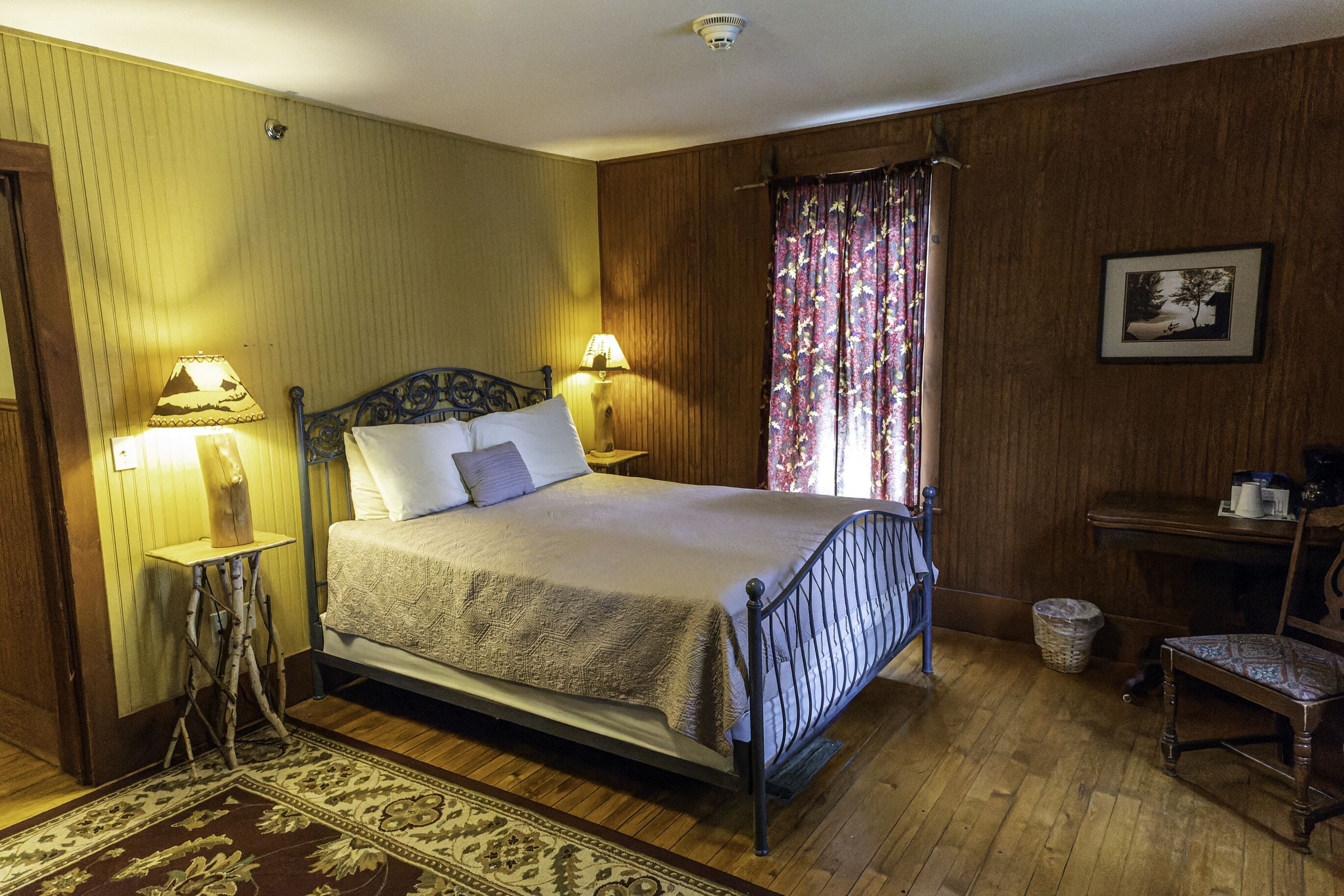 A traditional bedroom with a four-poster bed, two bedside lamps, a wooden floor, a patterned rug, and a small table with a chair.