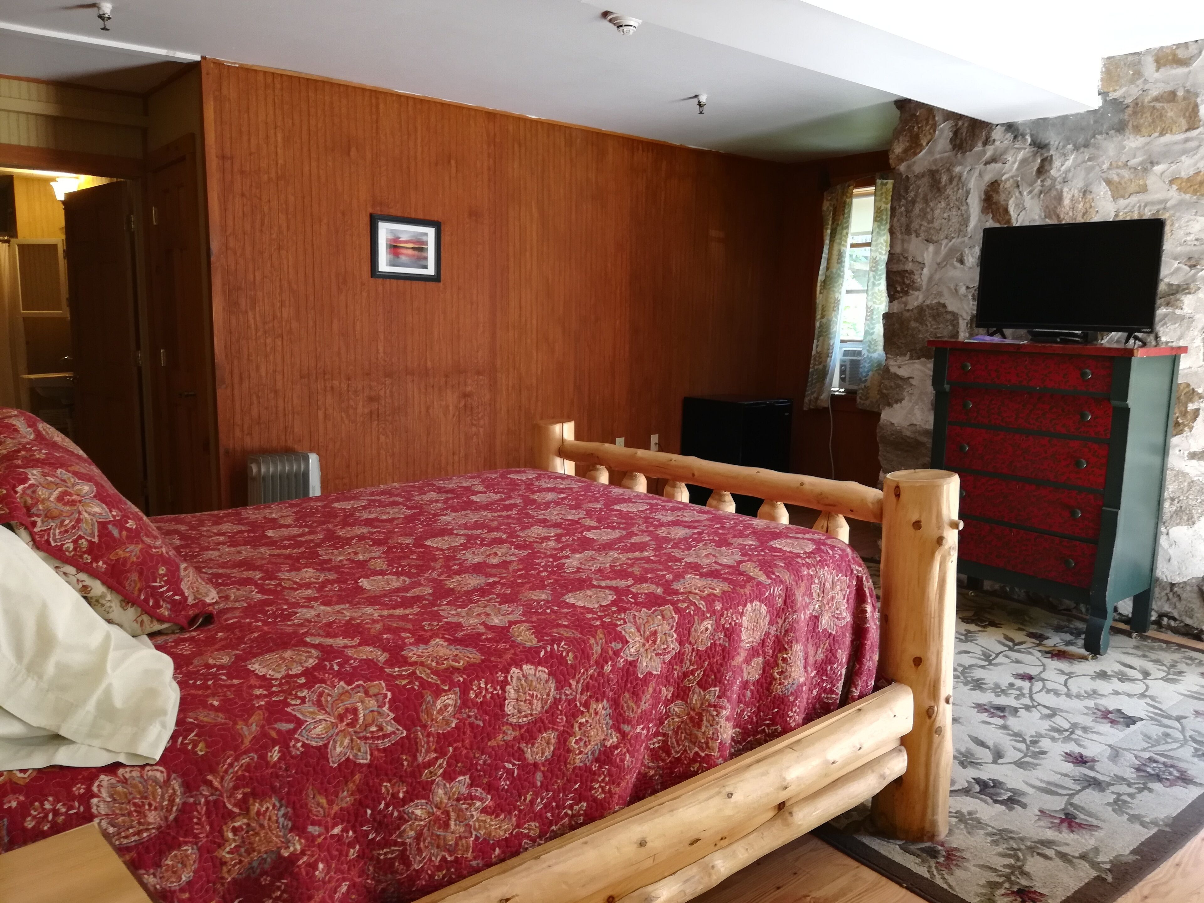 Une chambre à coucher avec un lit en bois, une couvre-lit aux motifs rouges et dorés, une commode en bois et un mur en pierre.