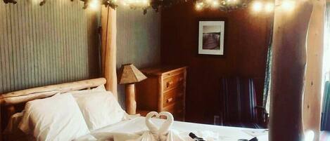 Habitación de hotel con cama, cabecera de madera, mesita de noche con lámpara, un cuadro enmarcado en la pared y una ventana con cortinas.