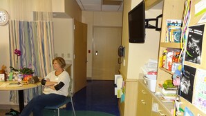 Une chambre d’hôpital avec un lit, une chaise, un bureau avec des effets personnels, une horloge et un tableau d’affichage où sont accrochés divers documents et photos.