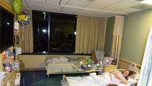 Une chambre d’hôpital avec deux lits, une table de chevet sur laquelle se trouvent divers objets, une fenêtre avec des rideaux et une vue sur l’extérieur.