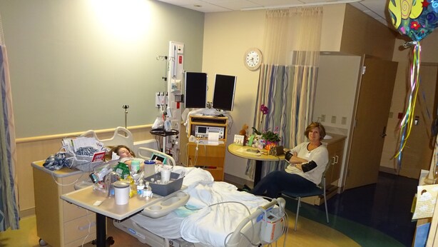 Une chambre d’hôpital avec un lit, du matériel médical, une table de chevet avec des effets personnels, une horloge au mur et un rideau.