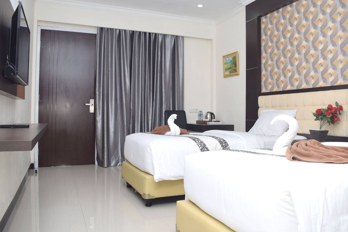 Habitación de hotel con dos camas, un televisor de pantalla plana montado en la pared, un ventanal con cortinas, un cuadro en la pared y una planta en maceta con flores rojas.
