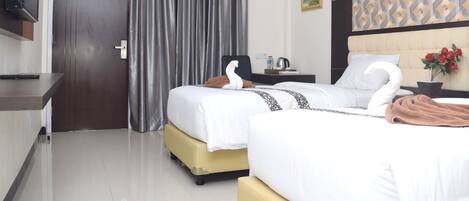 Habitación de hotel con dos camas, un televisor de pantalla plana montado en la pared, un ventanal con cortinas, un cuadro en la pared y una planta en maceta con flores rojas.
