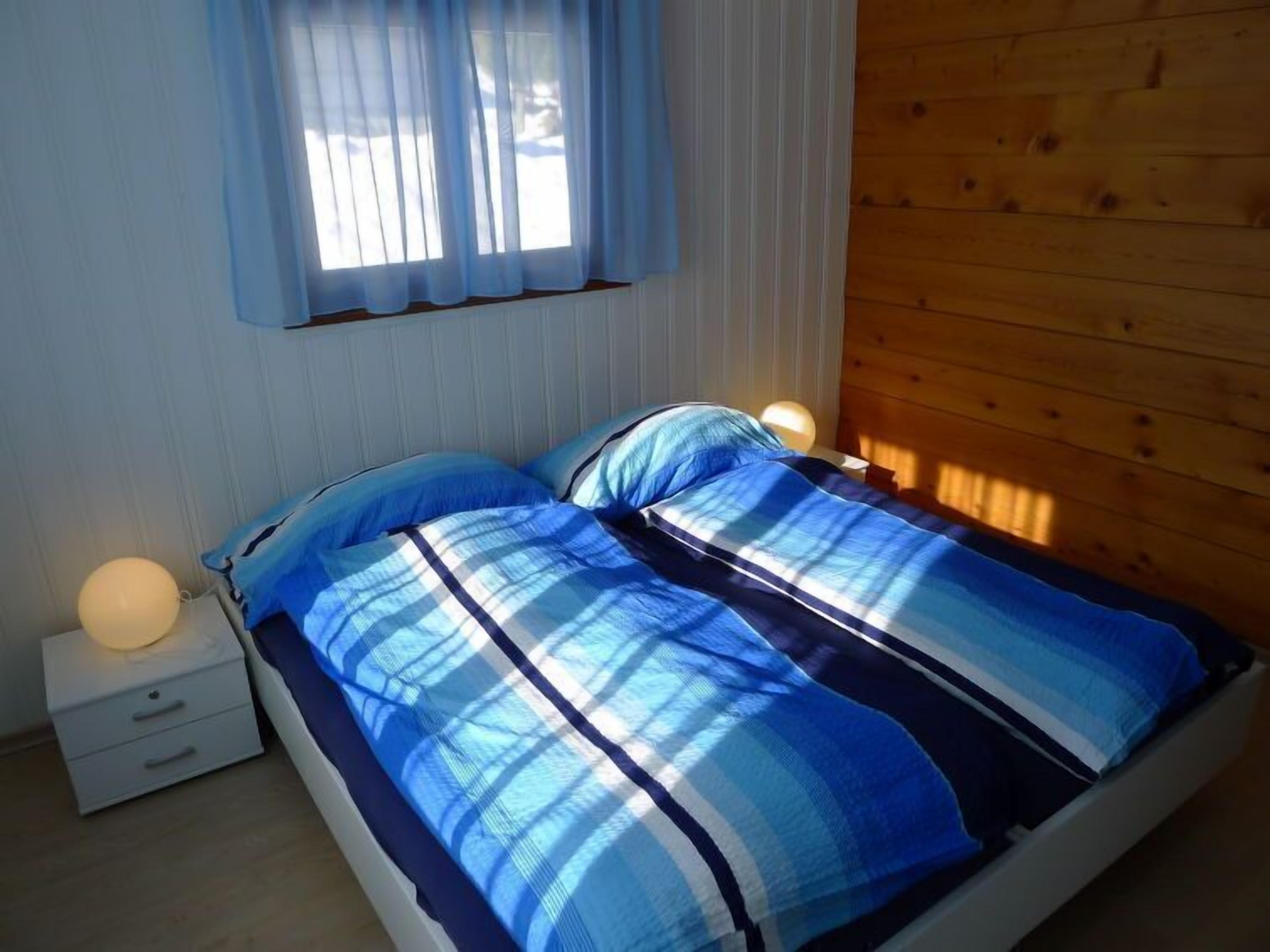 A bedroom with a bed, two bedside lamps, a nightstand, and a window with sheer curtains.