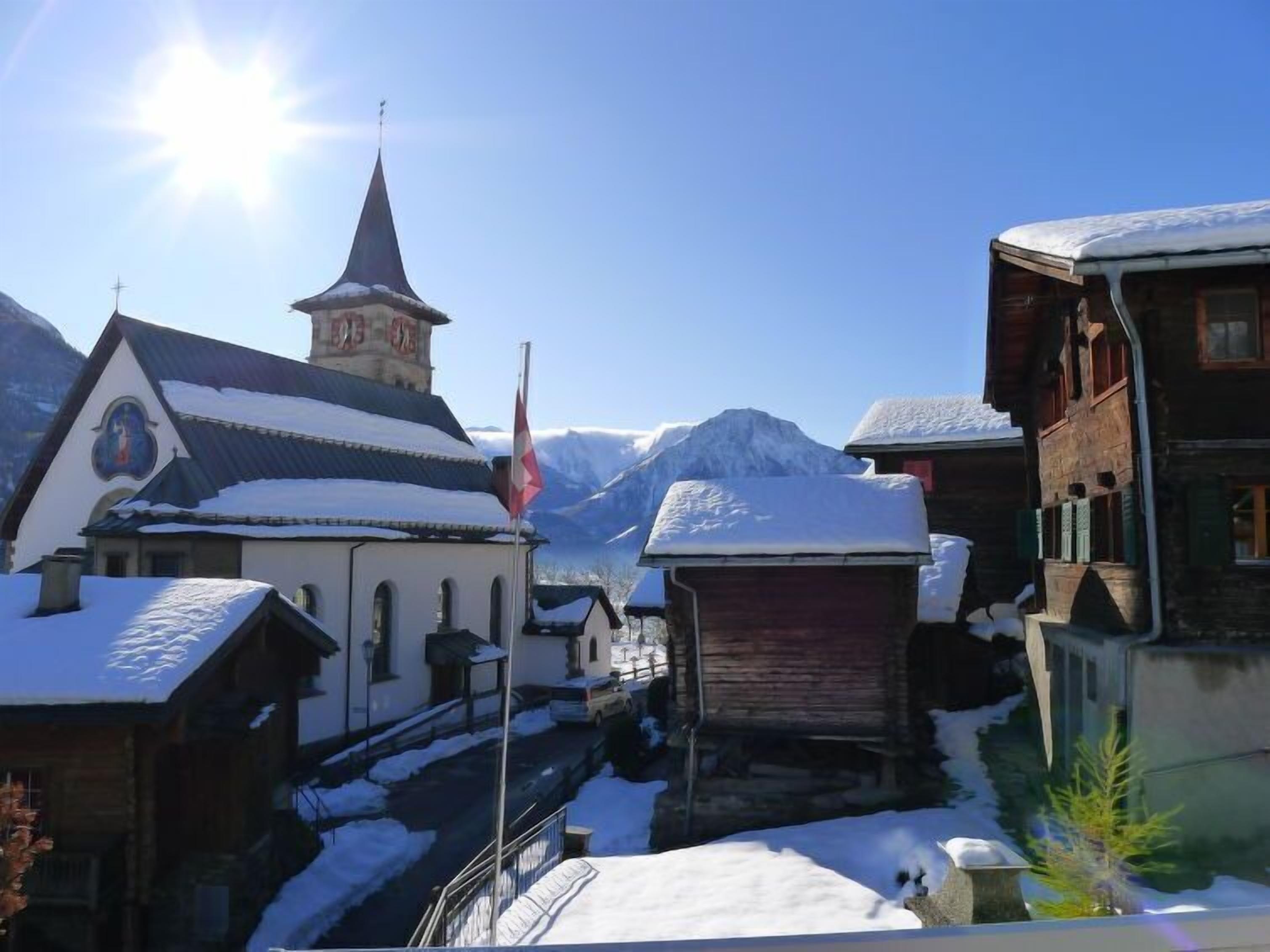 Un village enneigé, avec des bâtiments traditionnels, une église dotée d’un clocher et des montagnes en arrière-plan.