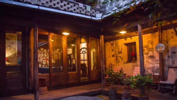 Um edifício rústico com portas e janelas de madeira, um banco de pedra e plantas em vasos.