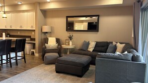Ein modernes Wohnzimmer mit einer Couch, einem Hocker und Sesseln, ein Essbereich mit Tisch und Stühlen sowie eine Küche mit Schränken und einer Lampe.