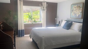 A bedroom with a large bed, a chandelier, a window with curtains, a plant, and a painting on the wall.