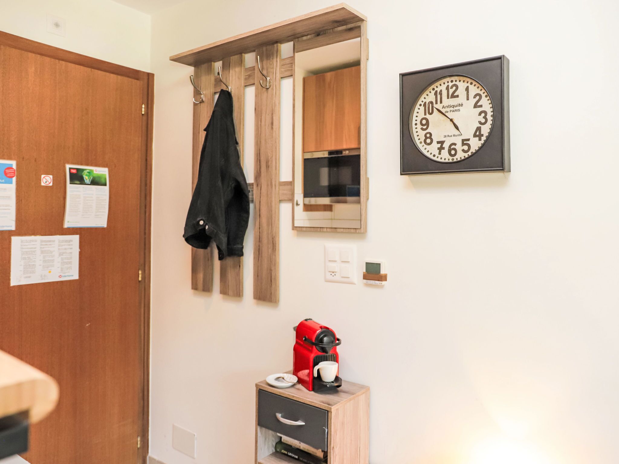 Ein Zimmer mit Holztür, einem Kleiderständer mit schwarzem Mantel, einer Wanduhr, einer kleinen Kaffeemaschine und einer Mikrowelle.