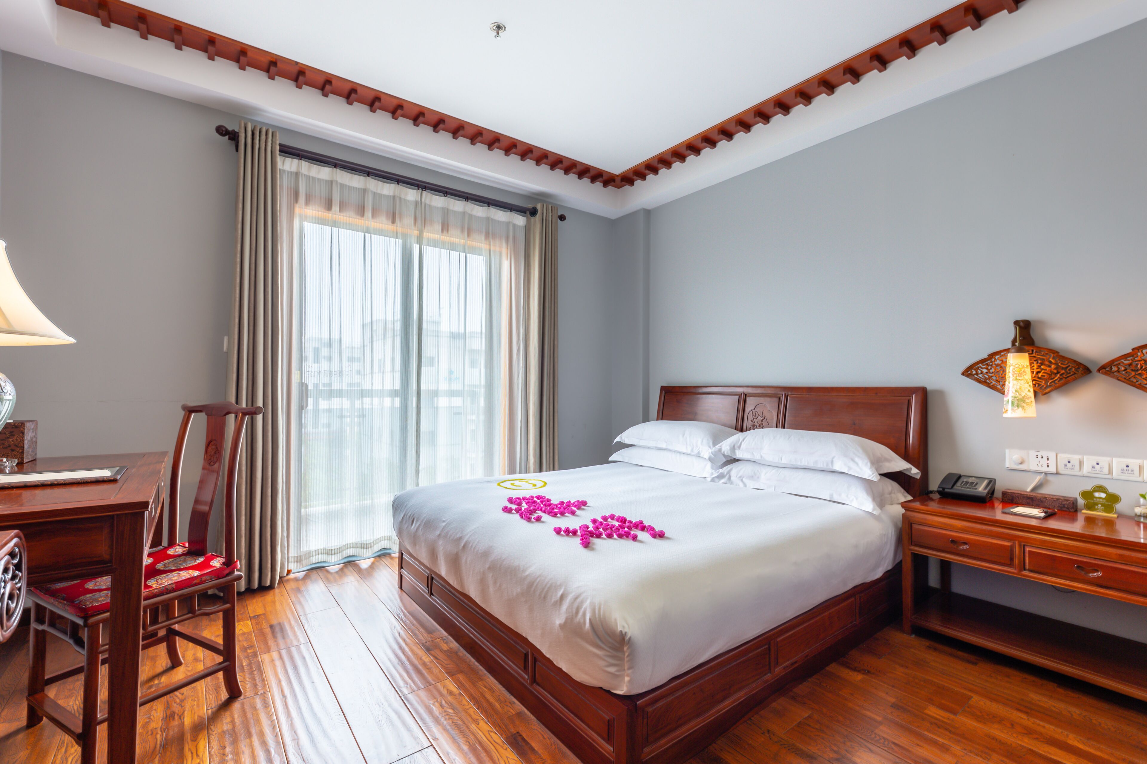 A hotel room with a wooden bed, a desk with a chair, a nightstand, and a window with curtains.
