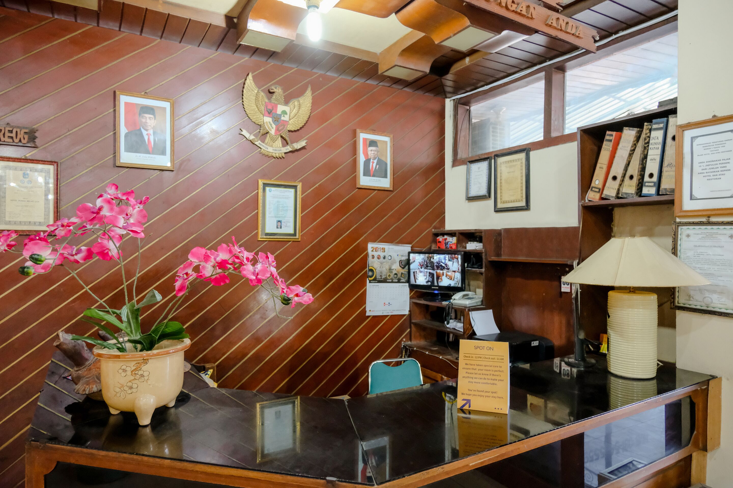 Un mostrador de recepción con fotografías enmarcadas, una orquídea en maceta y una pared de madera con un emblema heráldico.