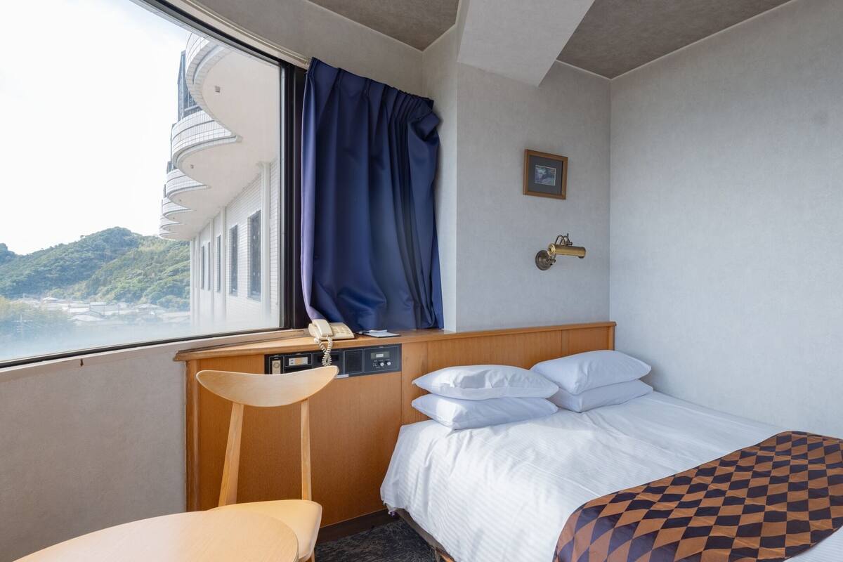 A hotel room with a bed, a desk with a telephone, a chair, and a window with curtains.