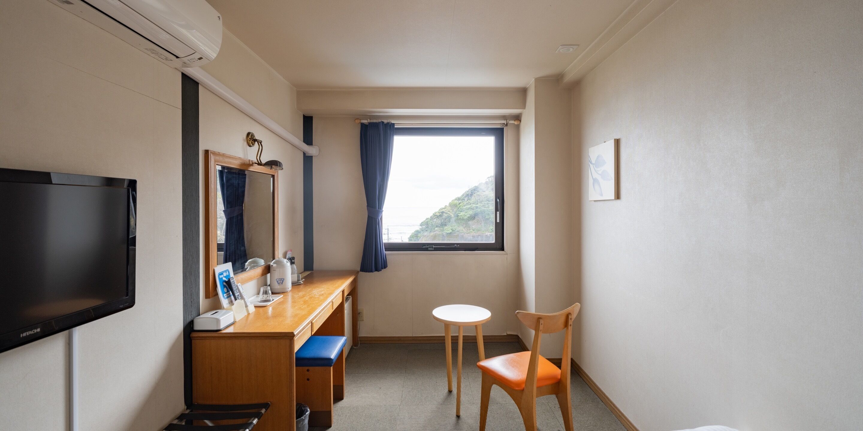 A hotel room with a flat-screen TV, a wooden desk with a mirror, a small round table, a wooden chair, and a window with a view of a mountain.