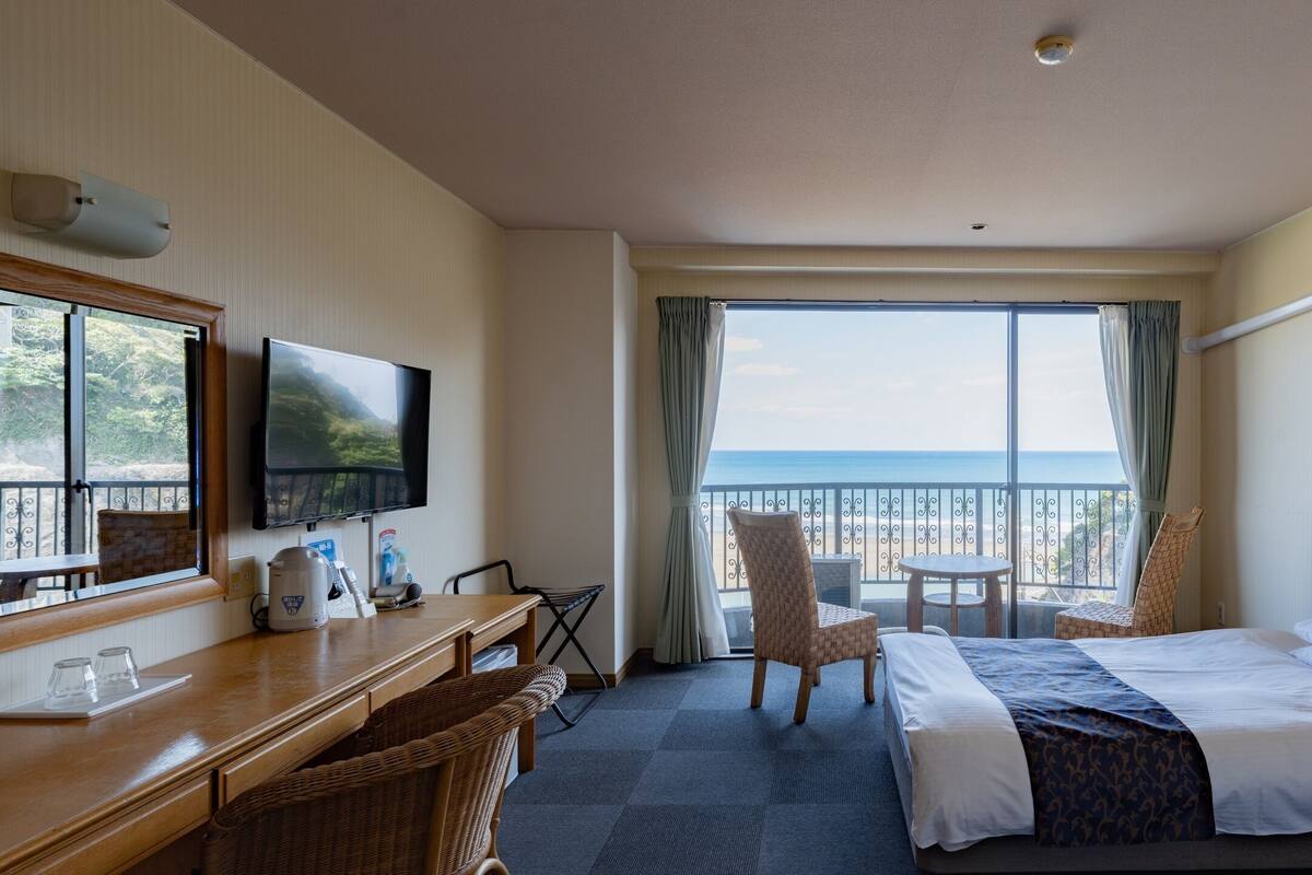 A hotel room with a bed, a desk with a chair, a TV, and a balcony with a view of the sea.