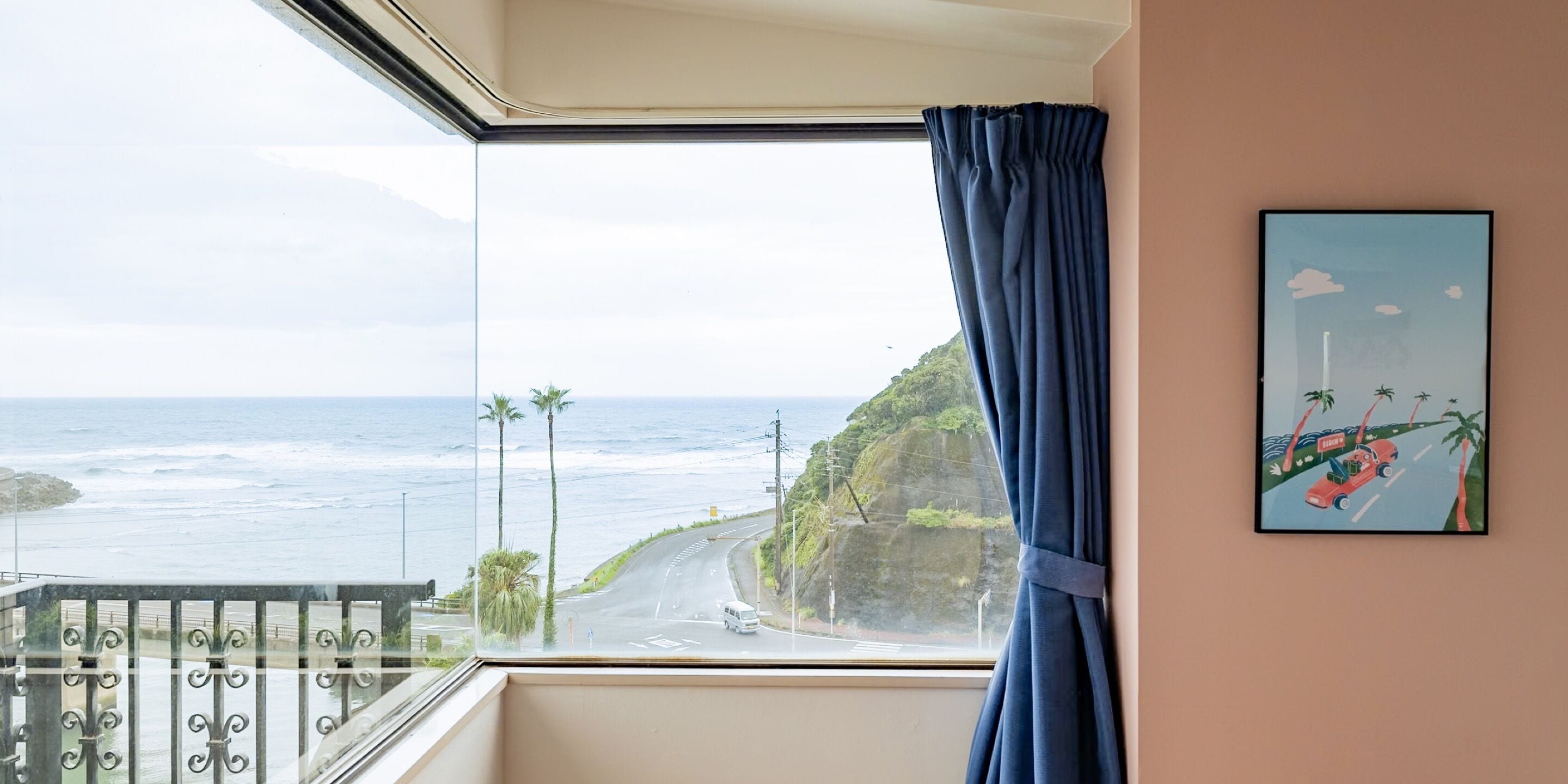 Una habitación con un ventanal que da a una carretera costera con palmeras y un acantilado.