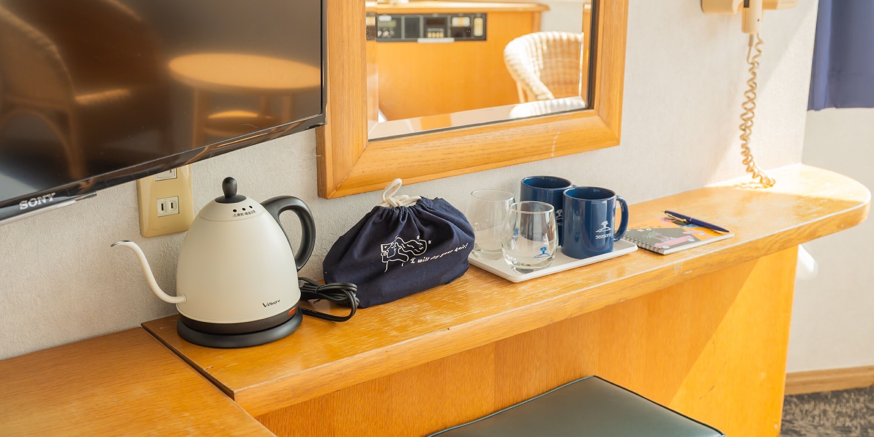 A hotel reception desk with a kettle, a black bag, two blue mugs, and a glass on a tray.
