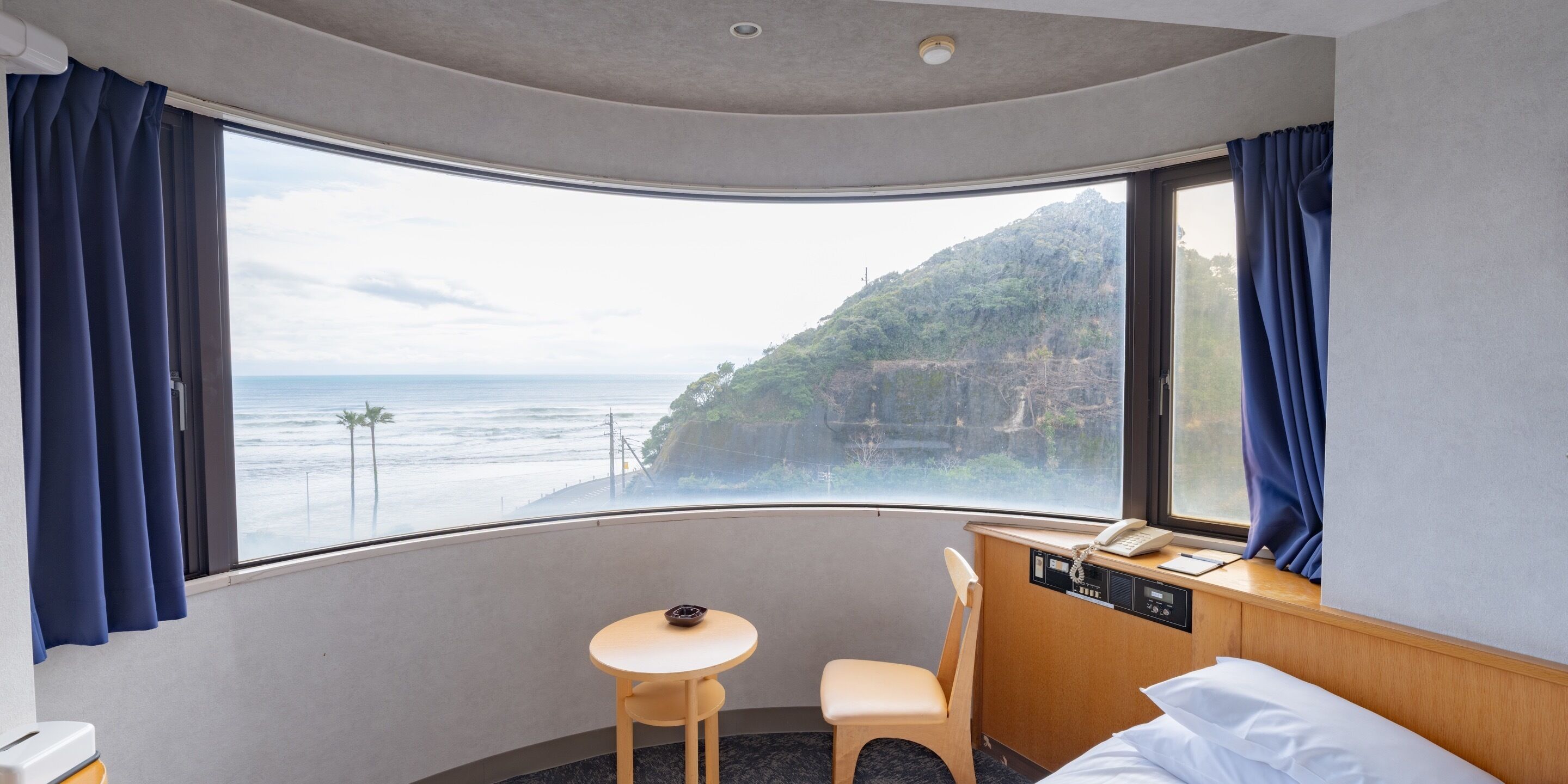A hotel room with a curved window offering a view of the ocean and a hillside. There is a bed, a small table, and a chair.