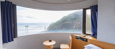 A hotel room with a curved window offering a view of the ocean and a hillside. There is a bed, a small table, and a chair.