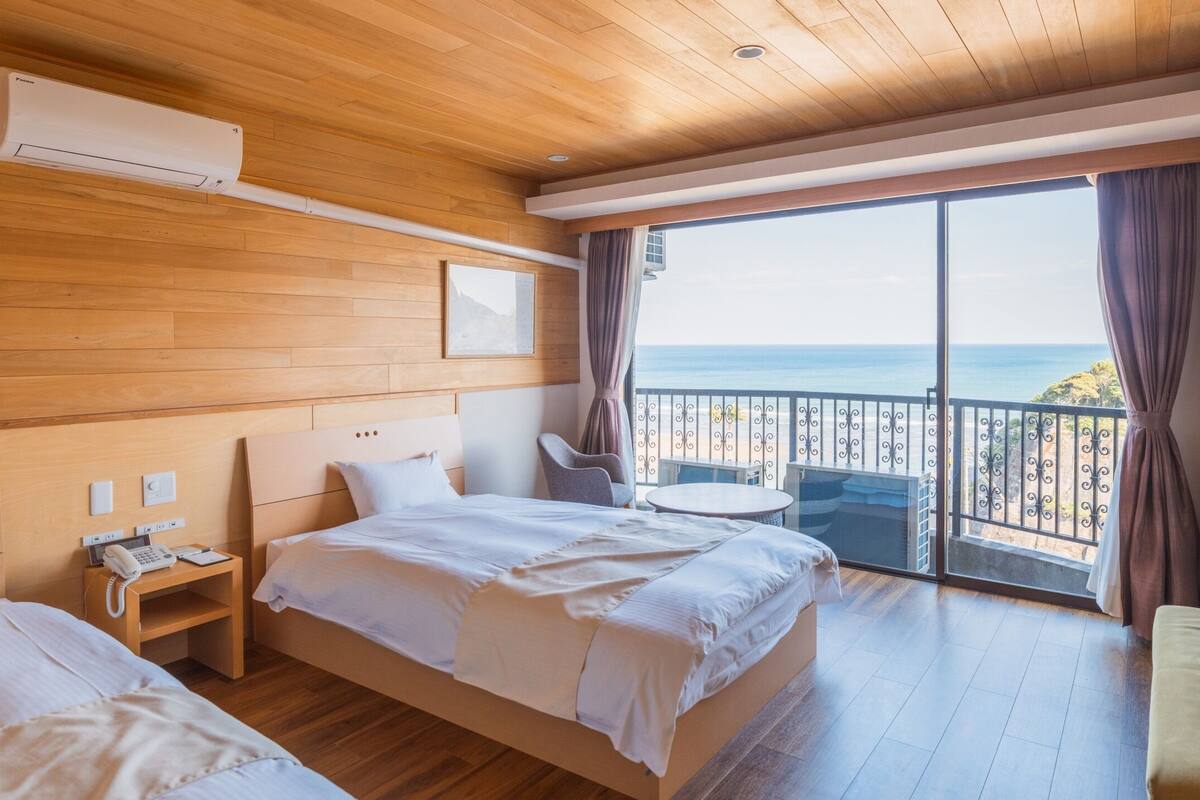 A hotel room with a wooden ceiling, a bed, a bedside table with a phone, a small round table with a chair, and a balcony with a view of the sea.