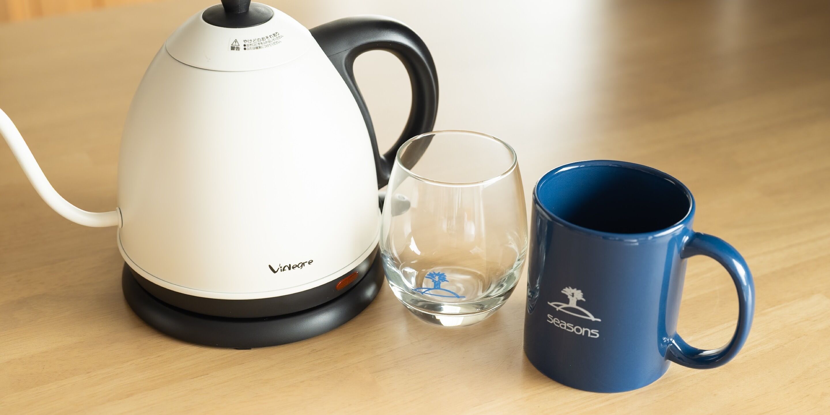 Une bouilloire électrique blanche, un verre transparent et une tasse bleue sur une table en bois.