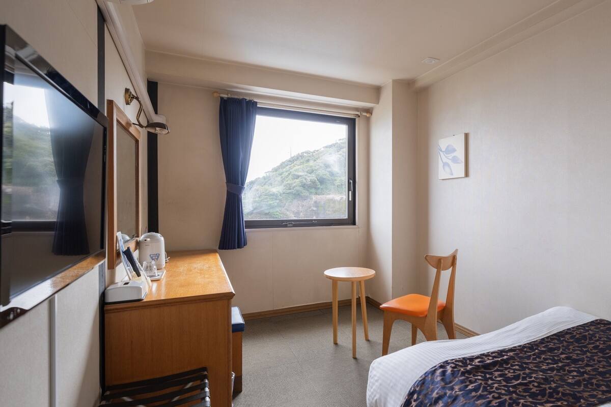 A hotel room with a bed, a desk with a chair, a television, and a window with a view of a hill.