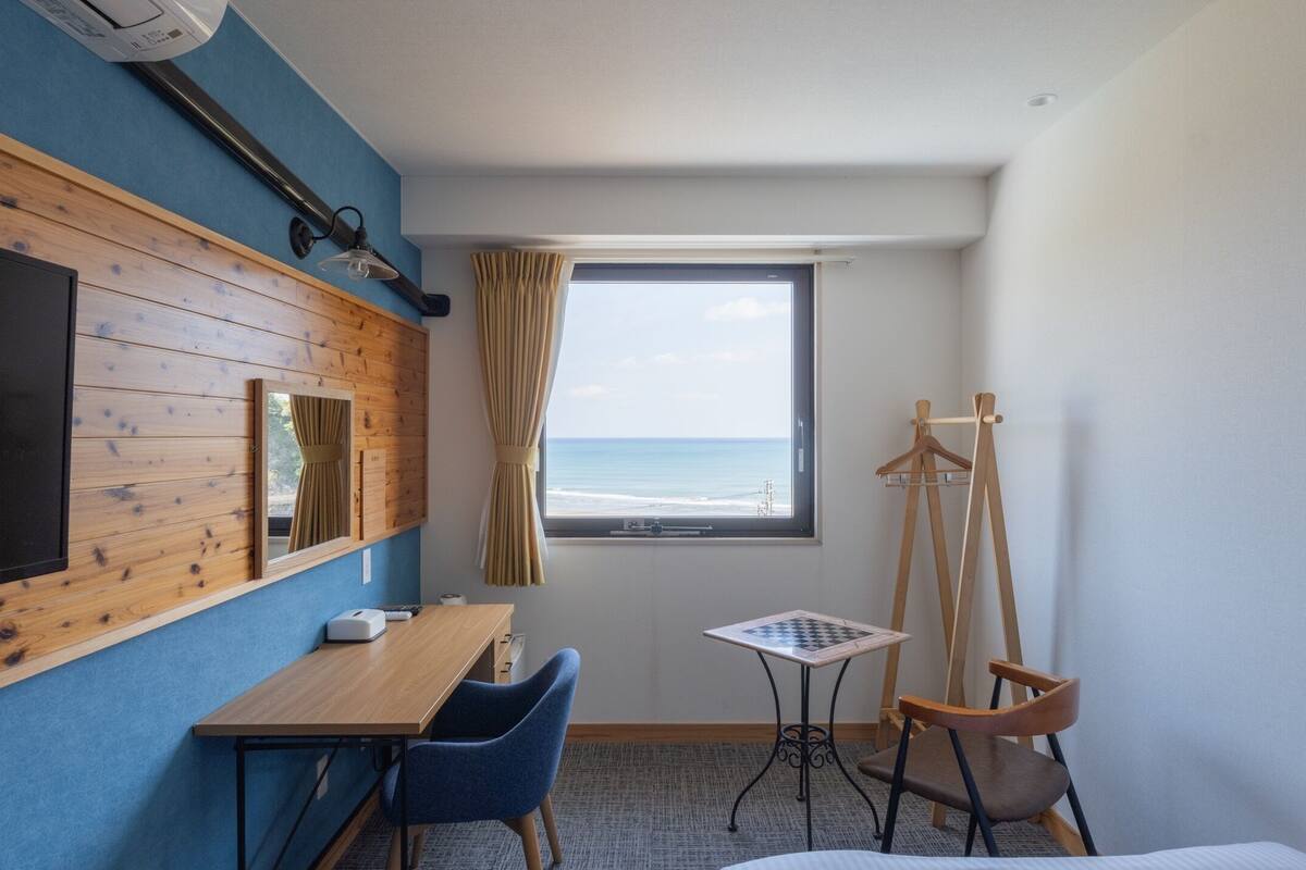 A hotel room with a wooden wall, a TV, a desk with a chair, a small table with a chessboard, a window with a view of the ocean, and a coat rack.