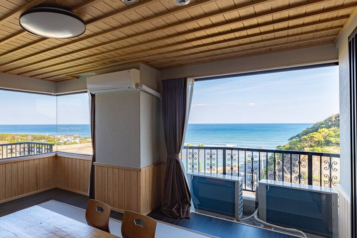 A room with a wooden ceiling, large windows with a view of the sea, a balcony with a railing, and a table with chairs.