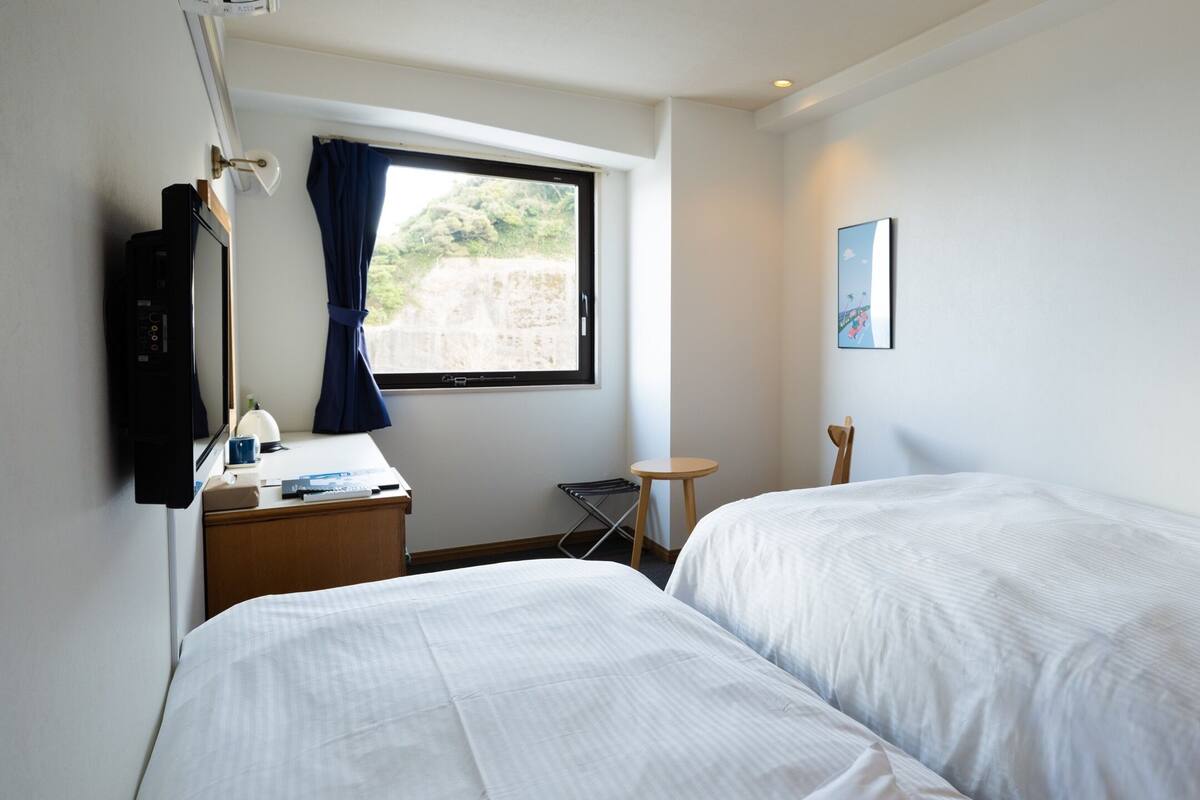 A hotel room with two beds, a desk with a lamp, a TV, a window with curtains, and a small table.