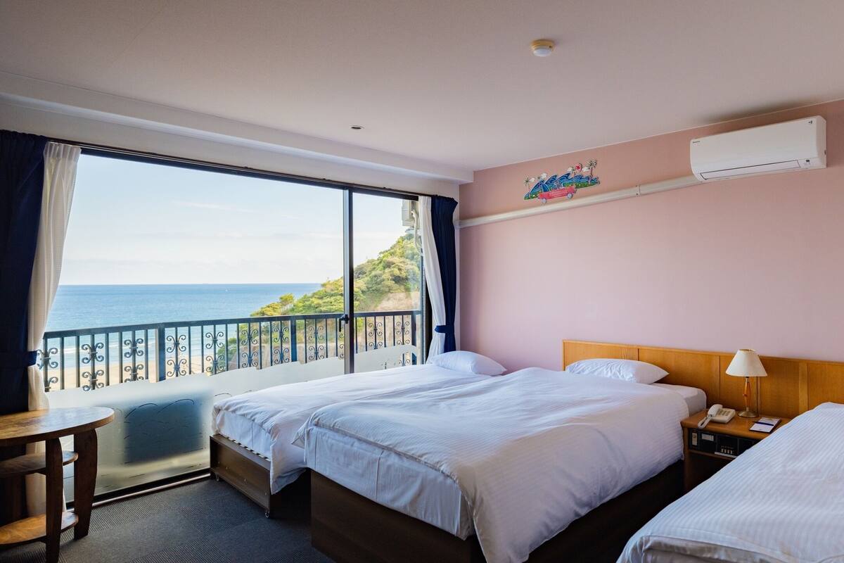 A hotel room with two beds, a bedside table, a lamp, and a view of the sea.