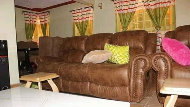 Un canapé en cuir marron agrémenté de plusieurs coussins, une petite table en bois et une table blanche se trouvent dans un salon doté de rideaux verts et d’une fenêtre.