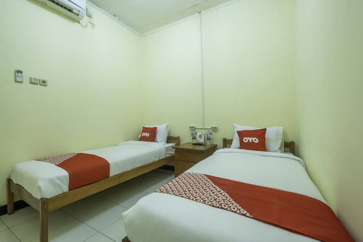 Ein Hotelzimmer mit zwei Einzelbetten, jeweils mit roter und weißer Bettdecke und passenden Kissen. Zwischen den Betten befindet sich ein Nachttisch mit einer Lampe und einem Telefon.
