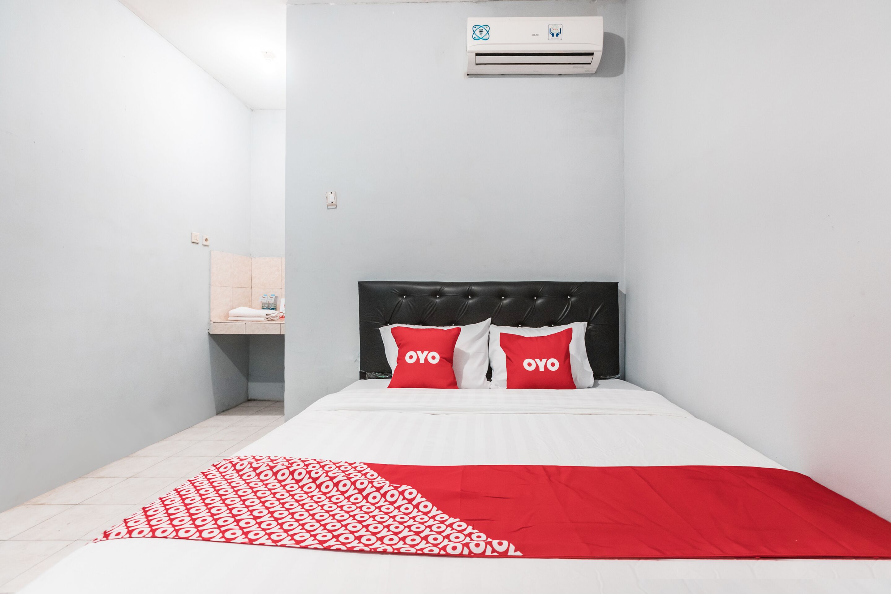 A hotel room with a bed, a red and white patterned blanket, a black headboard, and two red pillows with the word "OYO" on them.