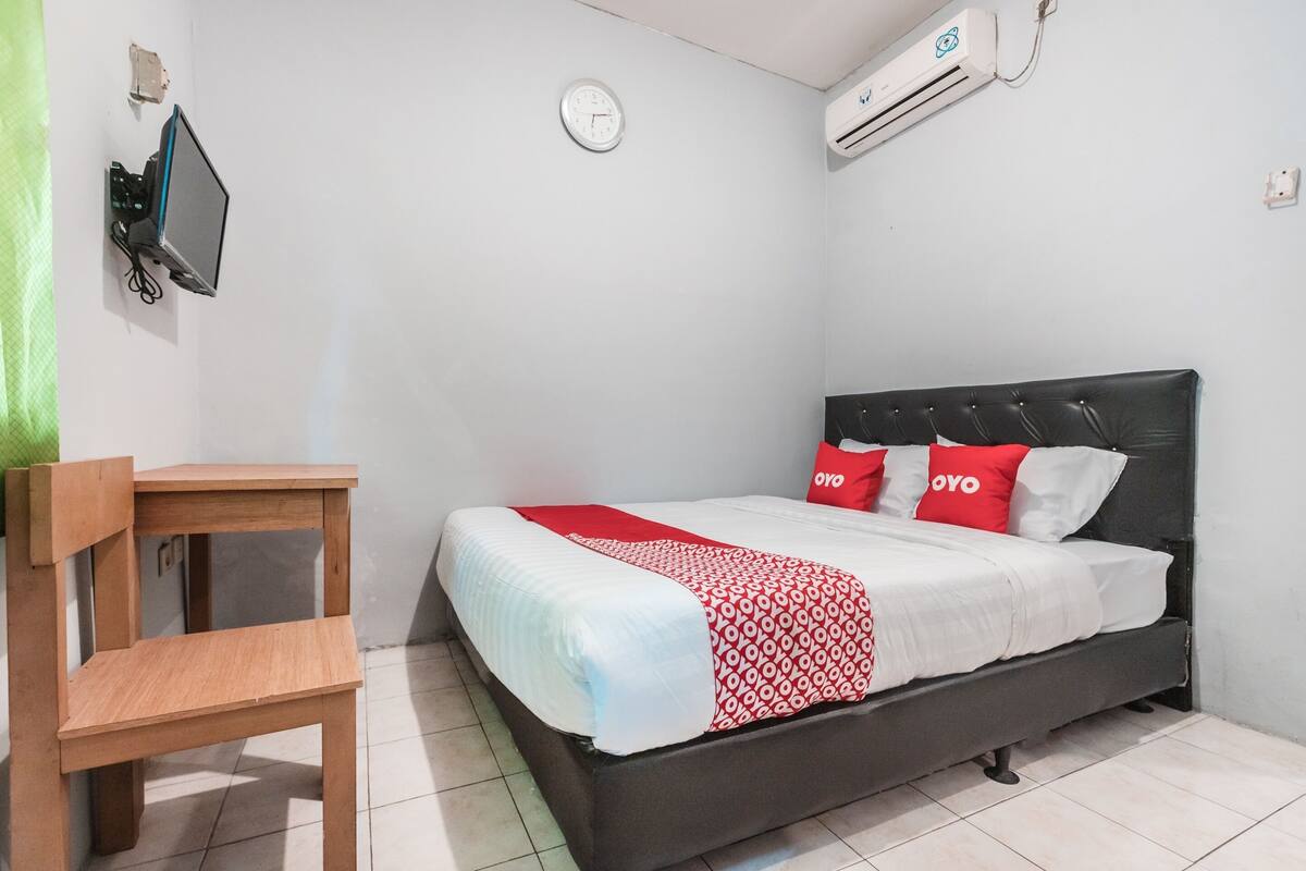 A bedroom with a bed, a wooden chair, a wall clock, and an air conditioner.