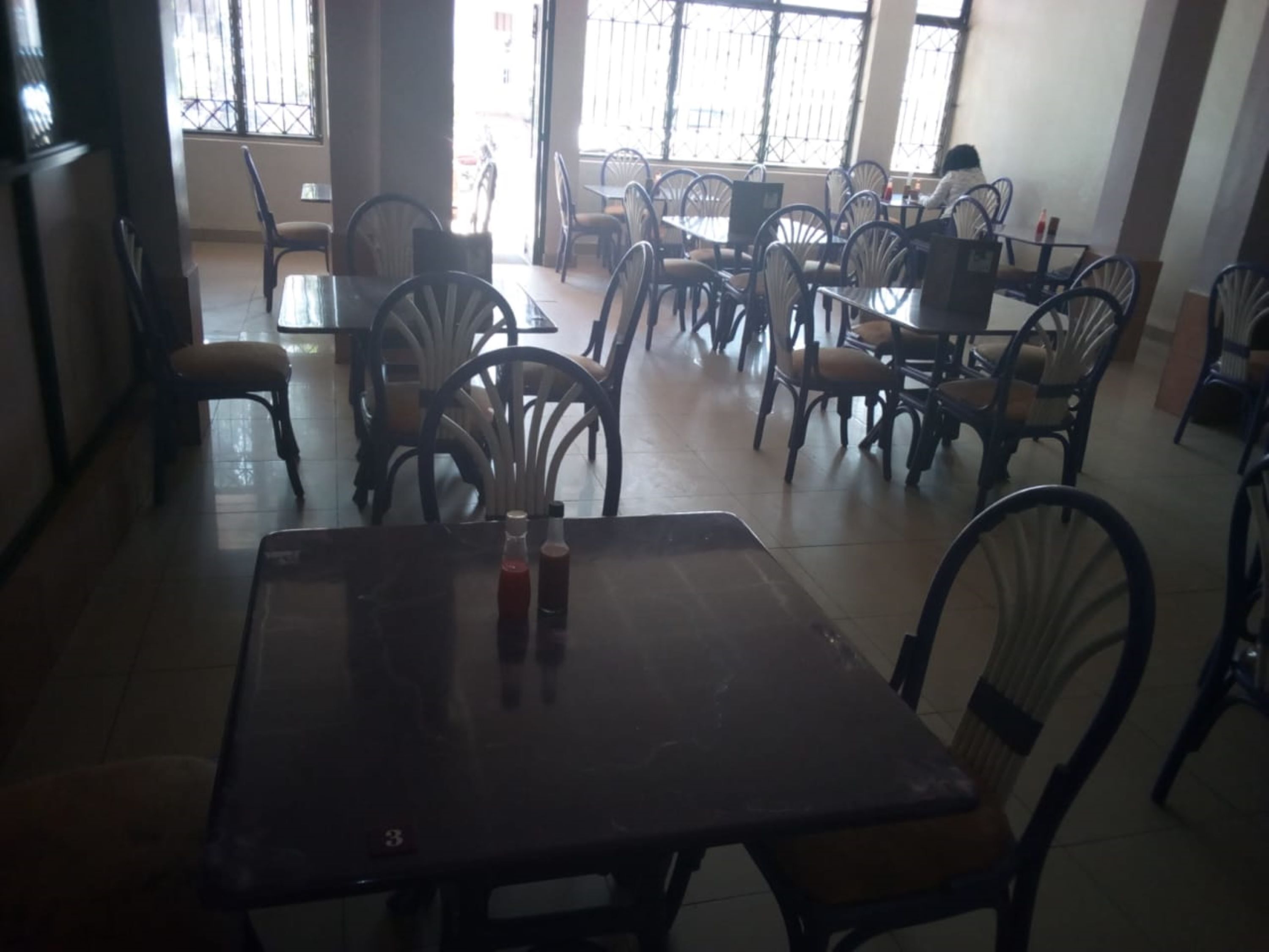 A dining area with multiple tables and chairs, a tiled floor, and large windows with metal bars.