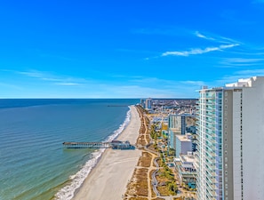 A coastal cityscape with a beach, a pier, and high-rise buildings.