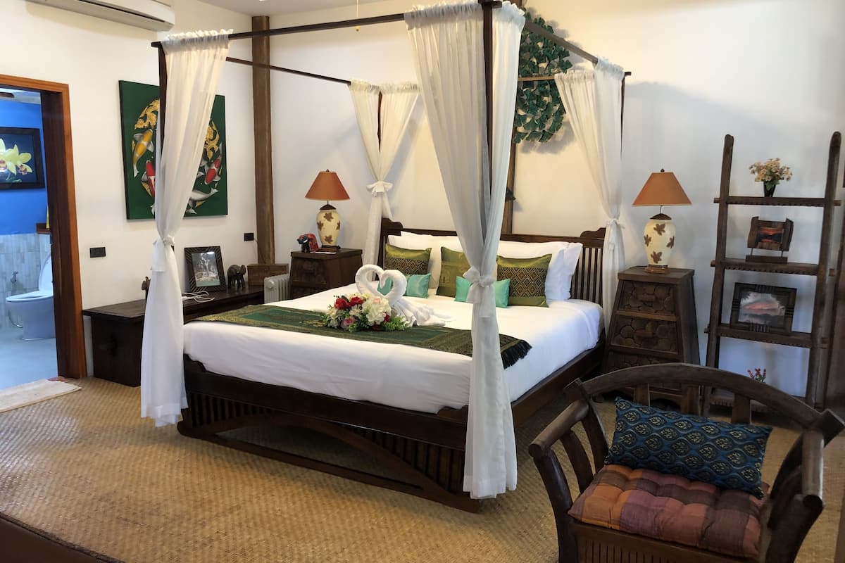 A four-poster bed with white linens, a bouquet of flowers, and a heart-shaped towel arrangement. There are two bedside tables with lamps, a wooden chair with a cushion, and a wooden ladder shelf with pictures.