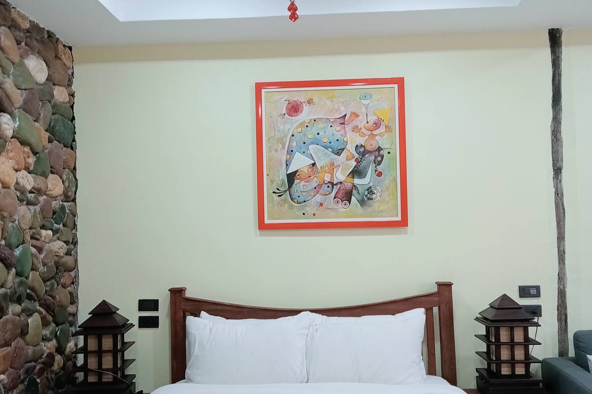 A bedroom with a bed, a ceiling fan, a stone wall, a painting, and a nightstand with a lantern.