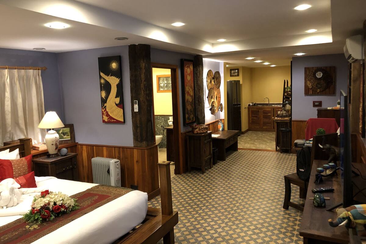 A hotel room with a bed, a desk, a chair, a small table, a lamp, and a bathroom visible through an open door.
