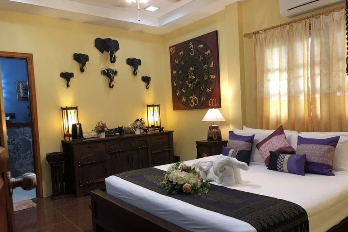 A hotel room with a large bed, a wooden dresser, a wall clock, and a decorative elephant head on the wall.
