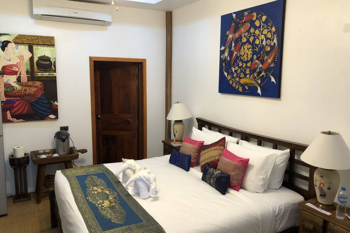 A hotel room with a bed, pillows, a wooden headboard, a painting, a wooden door, a table with a lamp, and a water bottle.