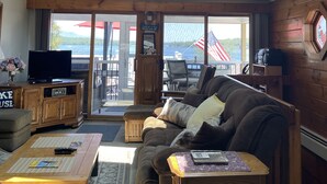 A spacious living room with a large wooden ceiling, a ceiling fan, a clock, a television, and a view of the outdoors through large sliding glass doors. - Weirs Beach - PRIME LOCATION!!!  (Weirs Beach)