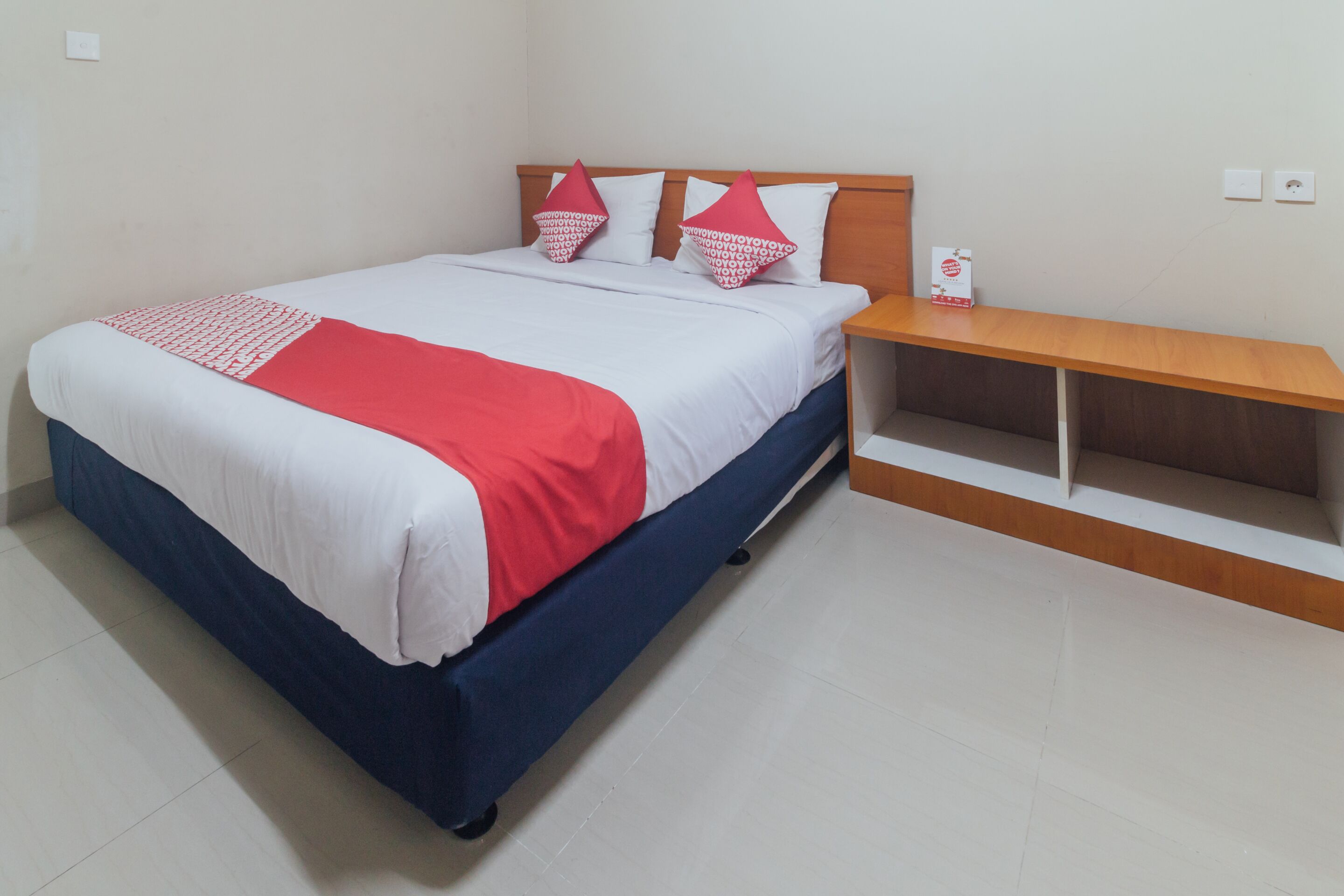 A neatly made bed with a red and white bedspread, two pillows, and a wooden nightstand with a box on it.
