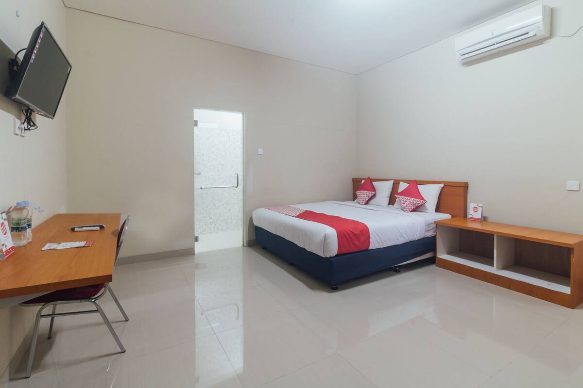 A hotel room with a bed, a desk, a chair, a TV, and an air conditioning unit.
