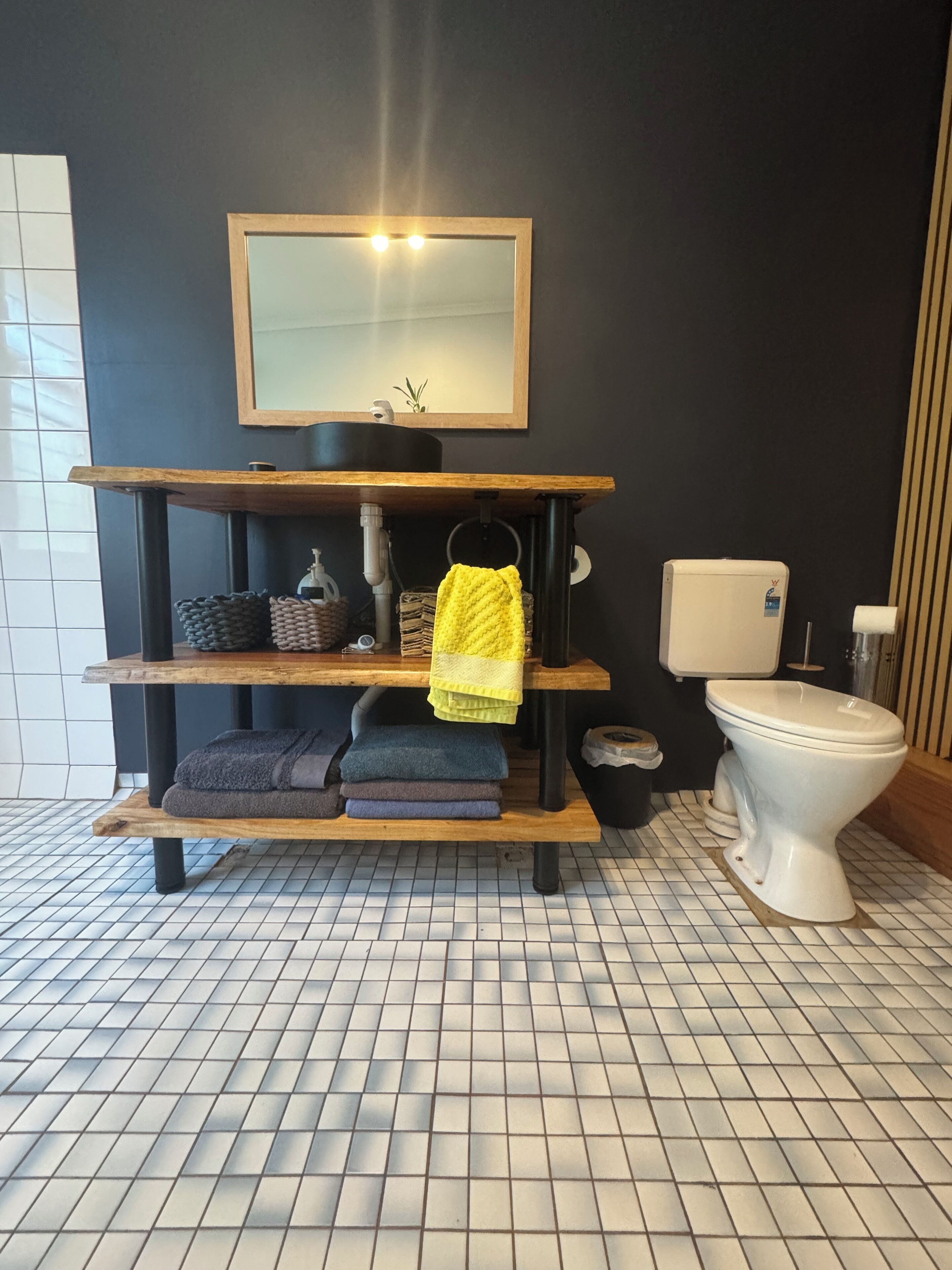 Een badkamer met betegelde vloer, een houten plank met handdoeken, een toilet en een spiegel boven de wastafel.