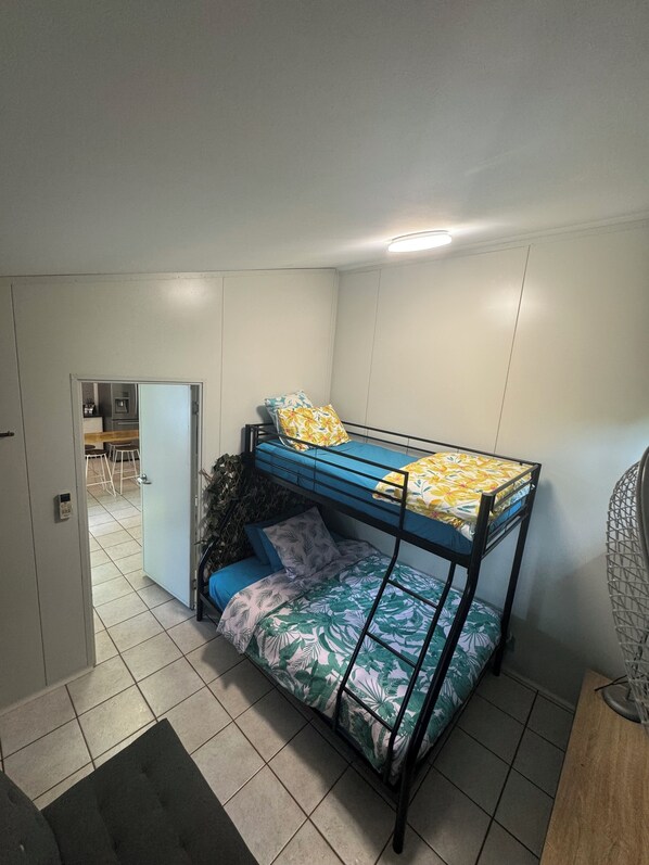 Une chambre avec un lit superposé, un petit bureau et une chaise, une porte donnant sur la cuisine et un sol carrelé.