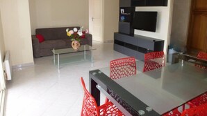 A modern living room with a glass coffee table, a sofa, a TV mounted on the wall, and a dining area with a glass table and red chairs. - APARTMENT T3 - Private parking - 300 m SEA & BEACH (Fréjus)
